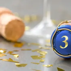 Sektkorken mit blauer Kappe und goldener "3" symbolisiert die Bedeutung des 3. Hochzeitstags. Goldkonfetti und ein weiterer Korken im Hintergrund deuten auf eine Feier hin.