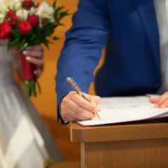 Ein Mann im blauen Anzug unterschreibt Dokumente, um standesamtlich zu heiraten. Die Braut hält einen roten und weißen Blumenstrauß.