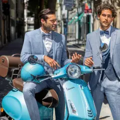 Zwei stilvolle Männer in hellblauen Anzügen, perfekt für einen Hochzeit Gast, posieren auf einer türkisfarbenen Vespa.