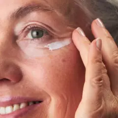 Eine Frau um die 60 trägt Augencreme auf. Ihre Haut zeigt natürliche Spuren des Alters, aber keine Pickel.