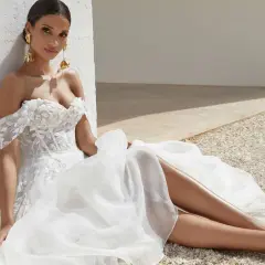 Eine Braut in einem weißen A-Linie Brautkleid mit floralen Applikationen sitzt lässig im Freien.
