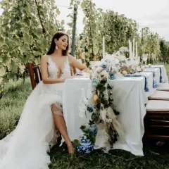 Braut in weißem Kleid sitzt am Tisch im Weinberg. Eine romantische Kulisse für eine weingut hochzeit.