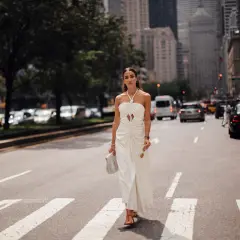 Eine Frau in einem weißen Kleid überquert eine Straße in New York. Sie trägt rote Sandalen und hält eine Sonnenbrille und eine Handtasche. Kleine Frauen sind hier nicht zu sehen.