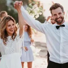 Glückliches Brautpaar feiert seine low budget hochzeit am Strand. Die Braut hält einen Blumenstrauß, der Bräutigam strahlt.