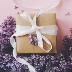 Ein Geschenkpaket mit weißem Band, umgeben von lila Flieder. Ein schönes Zeichen zum 13. Hochzeitstag.