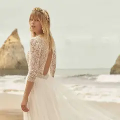 Eine Braut am Strand trägt ein luxuriöses Hochzeitskleid aus Spitze mit offenem Rücken und einem zarten Blumenkranz im Haar.