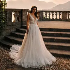 Eine Braut steht auf einer Steintreppe vor einer malerischen Bergkulisse. Ihr atemberaubendes Brautkleid mit zarter Spitze am Oberteil und einem fließenden Tüllrock ist ein Traum.
