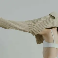 Eine Frau mit dunklem Haar trägt einen weißen BH. Ein beiger Strickpullover wird über ihren Kopf gezogen.