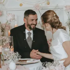 Ein glückliches Brautpaar bei ihrer Petersilienhochzeit, umgeben von zarten rosa Rosen und Schleierkraut. Sie halten Händchen und blicken sich liebevoll an.