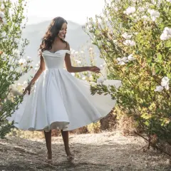 Glückliche Braut in einem schwingenden, knielangen Brautkleid mit schulterfreiem Ausschnitt, umgeben von blühenden Büschen.