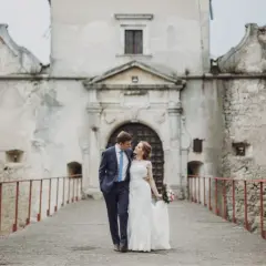 Ein Brautpaar spaziert über eine Brücke vor einer alten Burg. Die Szene erinnert an eine mittelalterliche Hochzeit.