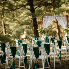 Reihen weißer Stühle mit grünen Schleifen schmücken den Weg zur Trauung. Eine romantische Hochzeit im Wald.