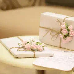 Zwei Geschenke, mit rosa Rosen und Juteband verziert, stehen auf einem Tisch. Ein Brief liegt daneben. Ein schönes Bild, das die Bedeutung des 14. Hochzeitstags unterstreicht.