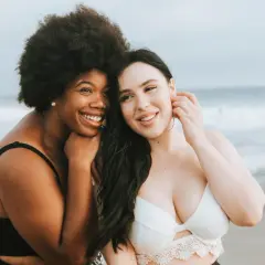 Zwei Frauen am Strand, eine mit Afro, die andere mit langen dunklen Haaren. Beide tragen Bikinis und lächeln. Ihre Brust und Bikinifigur sind gut sichtbar.