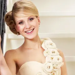 Lächelnde Frau in einem schulterfreien Kleid in Farbe ivory, verziert mit Stoffrosen. Sie lehnt an einem Geländer.