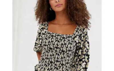 Frau mit lockigem Haar trägt ein schwarzes Oberteil mit Gänseblümchen-Muster und einem modischen carre ausschnitt.