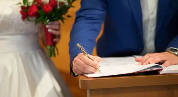 Ein Mann im blauen Anzug unterschreibt Dokumente, um standesamtlich zu heiraten. Die Braut hält einen roten und weißen Blumenstrauß.