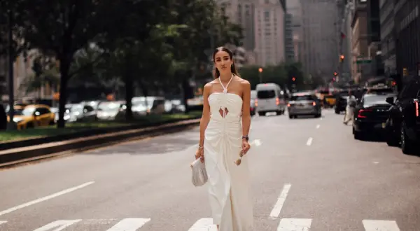Eine Frau in einem weißen Kleid überquert eine Straße in New York. Sie trägt rote Sandalen und hält eine Sonnenbrille und eine Handtasche. Kleine Frauen sind hier nicht zu sehen.