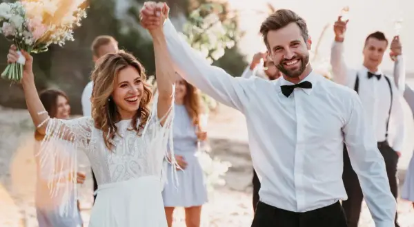 Glückliches Brautpaar feiert seine low budget hochzeit am Strand. Die Braut hält einen Blumenstrauß, der Bräutigam strahlt.