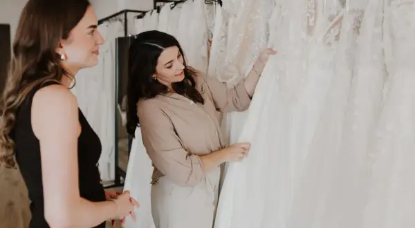 Zwei Frauen betrachten verschiedene Brautkleid-Stile in einem Geschäft. Eine Beraterin zeigt auf ein detailreiches Kleid.