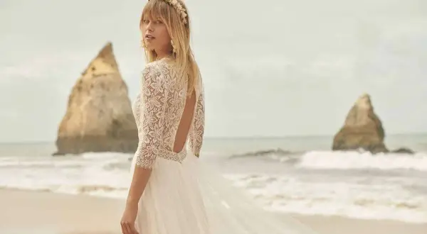 Eine Braut am Strand trägt ein luxuriöses Hochzeitskleid aus Spitze mit offenem Rücken und einem zarten Blumenkranz im Haar.