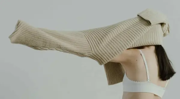 Eine Frau mit dunklem Haar trägt einen weißen BH. Ein beiger Strickpullover wird über ihren Kopf gezogen.