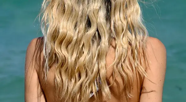 Blondes Haar mit Beach Waves im Wind, Schultern und Nacken einer Frau vor türkisblauem Wasser.