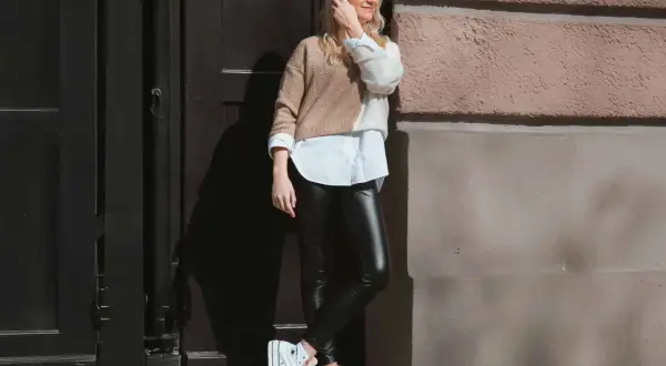 Stylische Frau mit blonden Haaren, die schwarze Lederhose kombiniert mit einem weißen Hemd und einem zweifarbigen Pullover trägt. Weiße Plateau-Sneaker runden den Look ab.