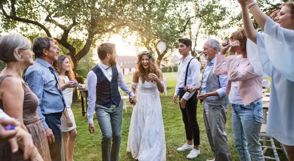 Brautpaar feiert mit Familie und Freunden. Konfetti fliegt, die Stimmung ist ausgelassen. Eine fröhliche Hochzeit, bei der die Farben der Kleidung und des Konfettis im Sonnenlicht leuchten.