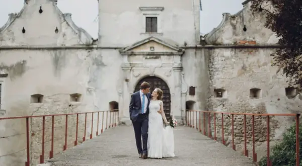 Ein Brautpaar spaziert über eine Brücke vor einer alten Burg. Die Szene erinnert an eine mittelalterliche Hochzeit.