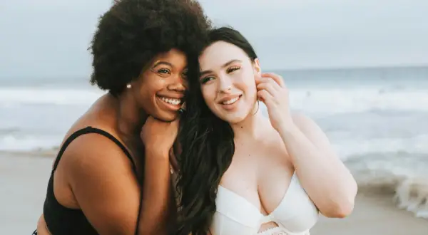 Zwei Frauen am Strand, eine mit Afro, die andere mit langen dunklen Haaren. Beide tragen Bikinis und lächeln. Ihre Brust und Bikinifigur sind gut sichtbar.