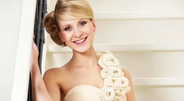 Lächelnde Frau in einem schulterfreien Kleid in Farbe ivory, verziert mit Stoffrosen. Sie lehnt an einem Geländer.