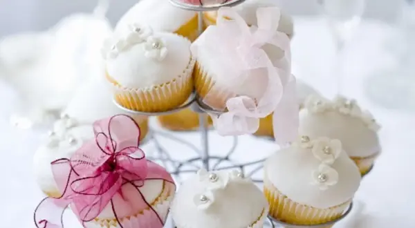 Zarte Muffins mit weißer Glasur, Blüten und Perlen, dekoriert mit rosa Bändern, perfekt für eine Hochzeit.