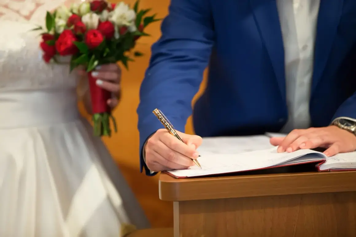 Ein Mann im blauen Anzug unterschreibt Dokumente, um standesamtlich zu heiraten. Die Braut hält einen roten und weißen Blumenstrauß.