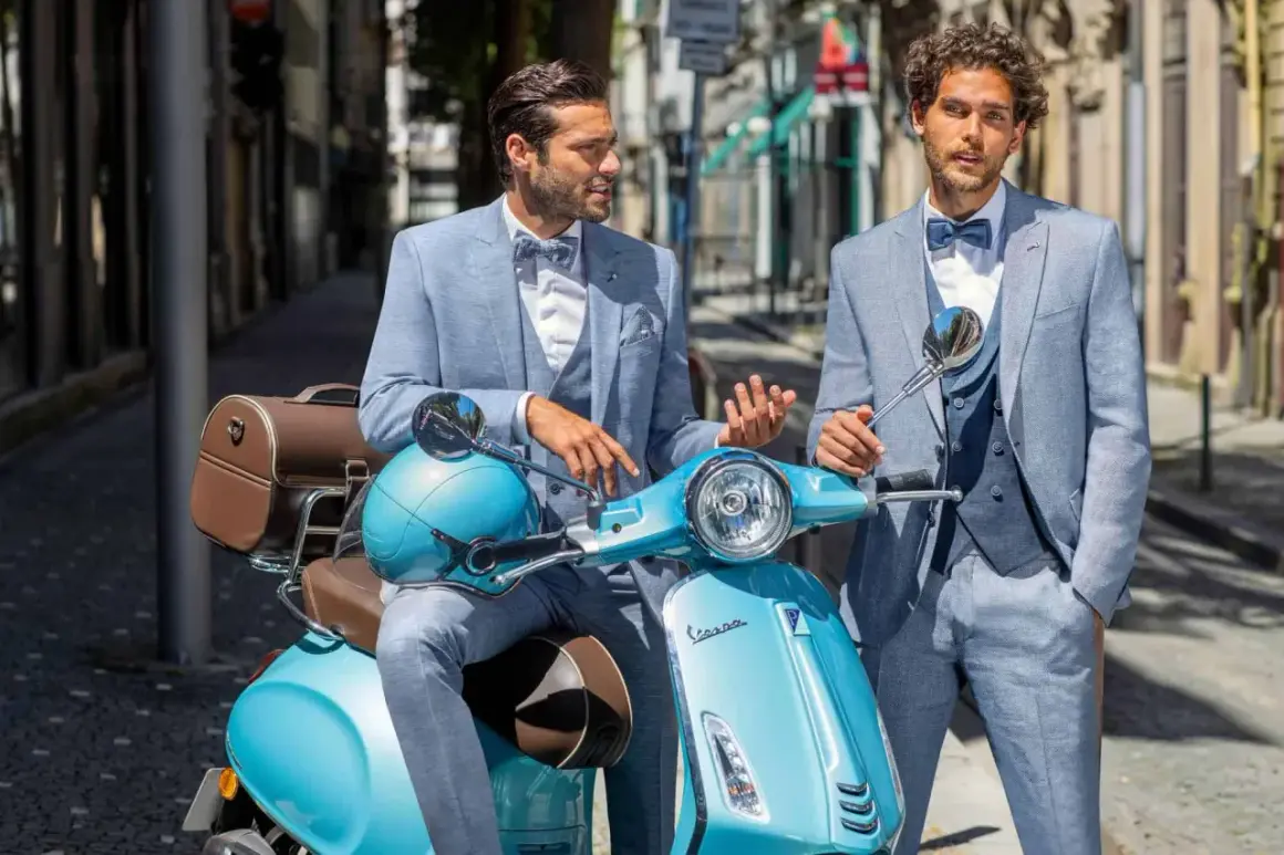 Zwei stilvolle Männer in hellblauen Anzügen, perfekt für einen Hochzeit Gast, posieren auf einer türkisfarbenen Vespa.