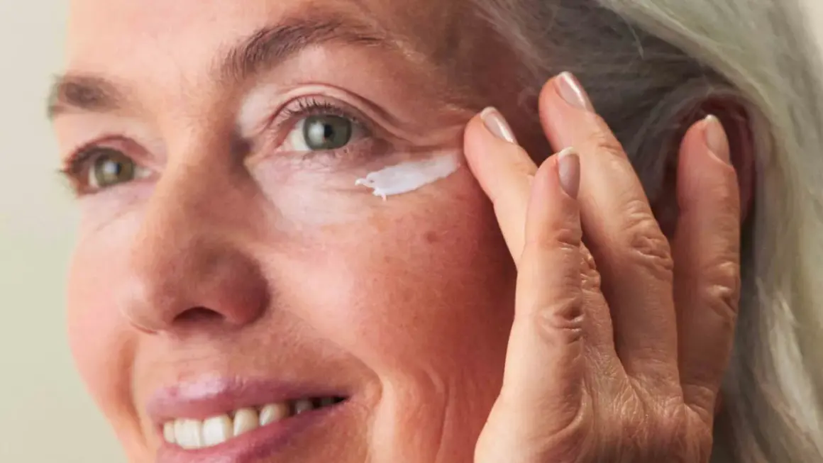 Eine Frau um die 60 trägt Augencreme auf. Ihre Haut zeigt natürliche Spuren des Alters, aber keine Pickel.