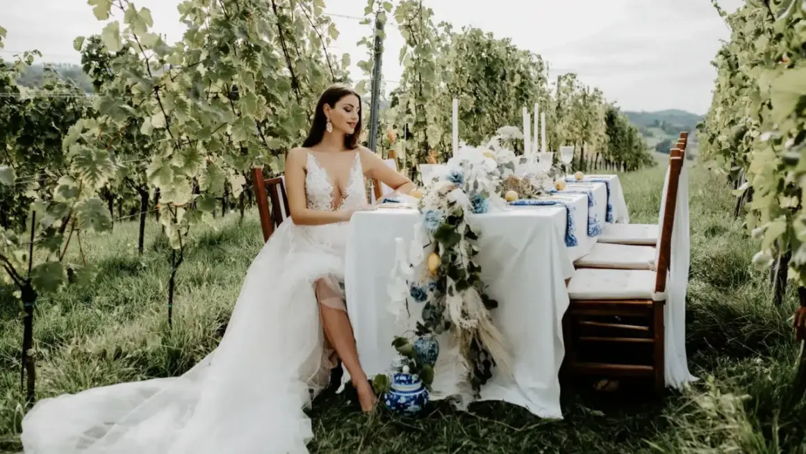 Braut in weißem Kleid sitzt am Tisch im Weinberg. Eine romantische Kulisse für eine weingut hochzeit.