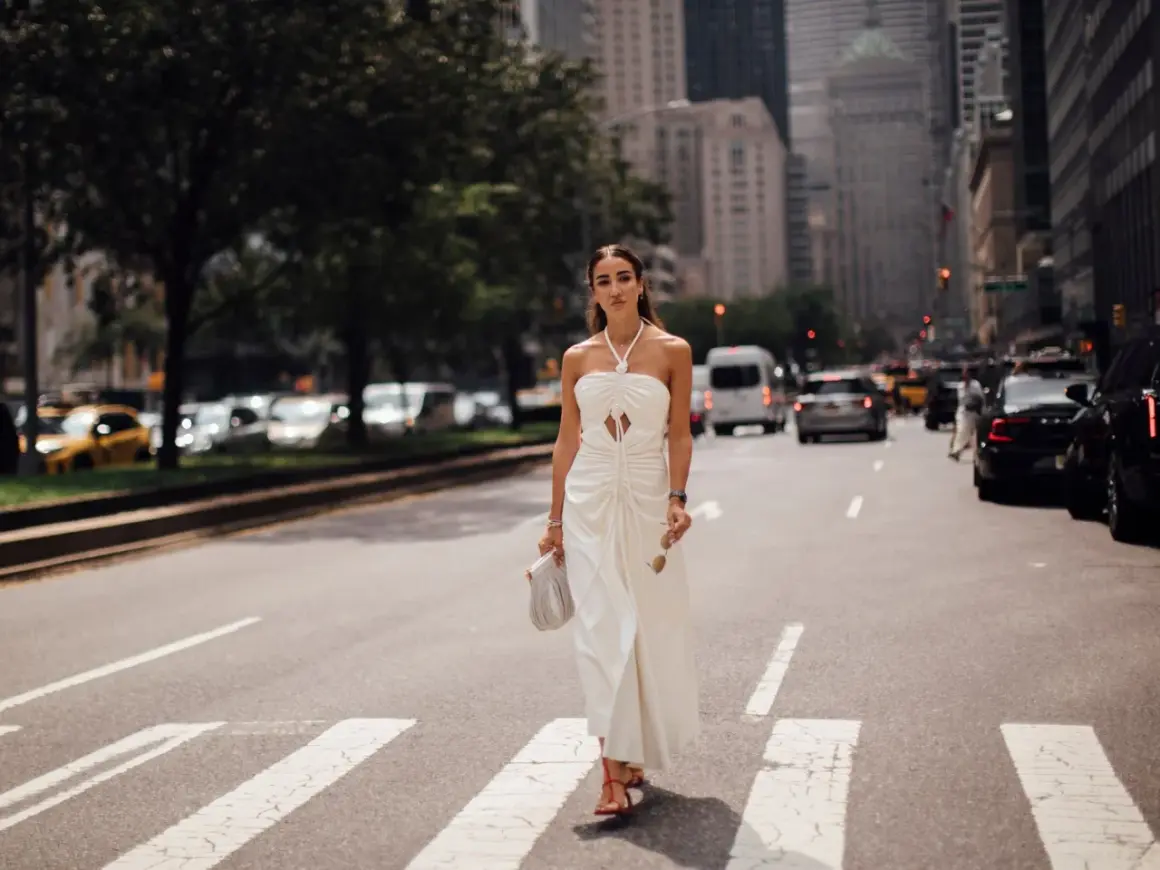 Eine Frau in einem weißen Kleid überquert eine Straße in New York. Sie trägt rote Sandalen und hält eine Sonnenbrille und eine Handtasche. Kleine Frauen sind hier nicht zu sehen.