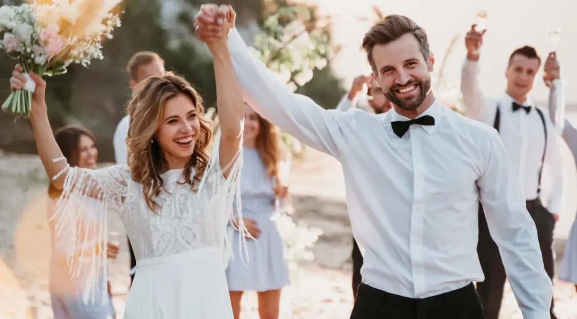 Glückliches Brautpaar feiert seine low budget hochzeit am Strand. Die Braut hält einen Blumenstrauß, der Bräutigam strahlt.