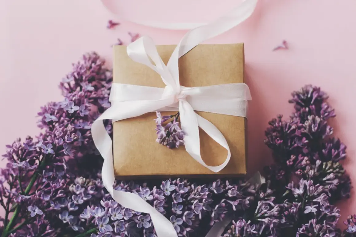 Ein Geschenkpaket mit weißem Band, umgeben von lila Flieder. Ein schönes Zeichen zum 13. Hochzeitstag.