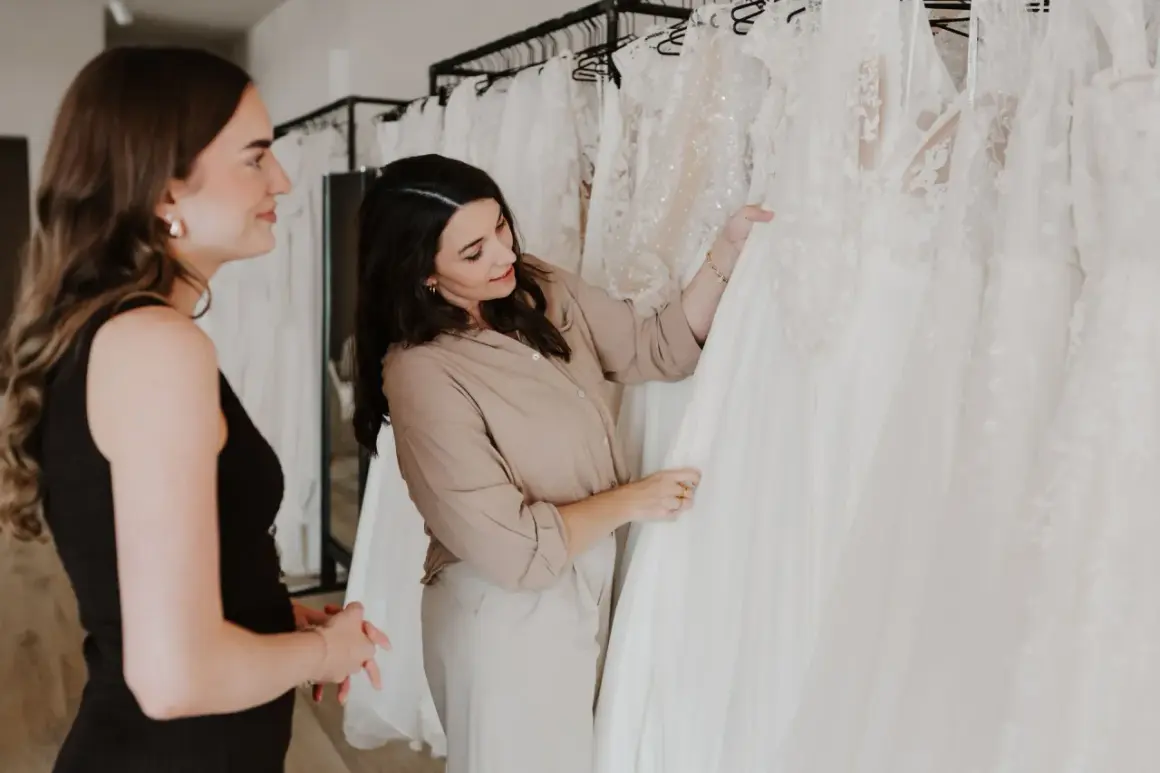 Zwei Frauen betrachten verschiedene Brautkleid-Stile in einem Geschäft. Eine Beraterin zeigt auf ein detailreiches Kleid.