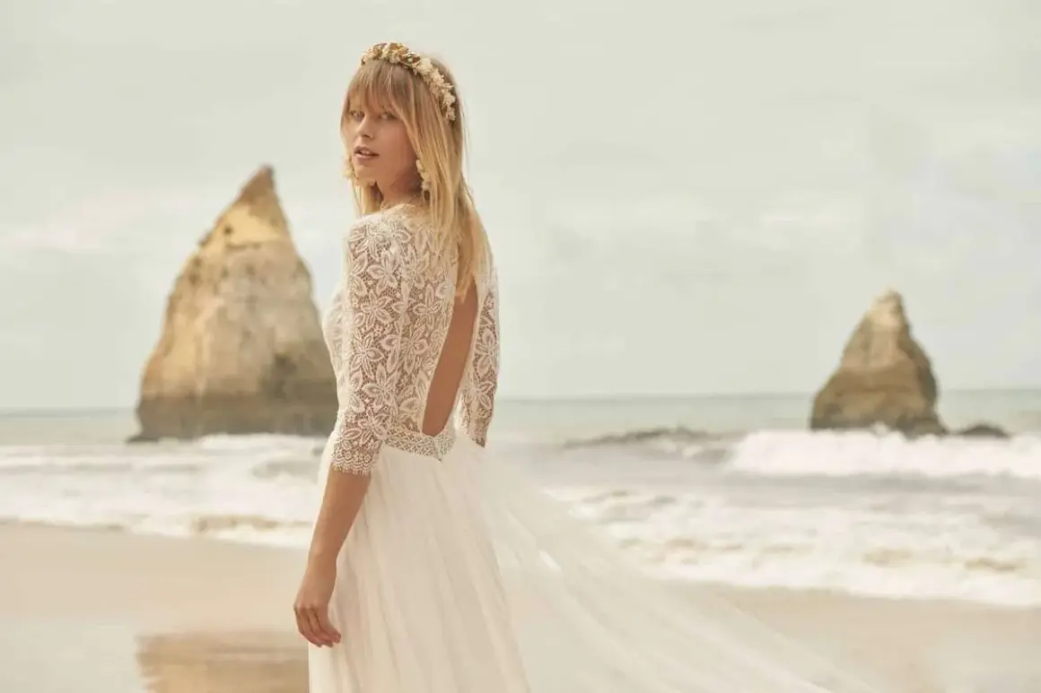 Eine Braut am Strand trägt ein luxuriöses Hochzeitskleid aus Spitze mit offenem Rücken und einem zarten Blumenkranz im Haar.