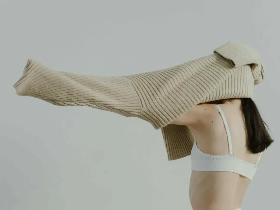 Eine Frau mit dunklem Haar trägt einen weißen BH. Ein beiger Strickpullover wird über ihren Kopf gezogen.