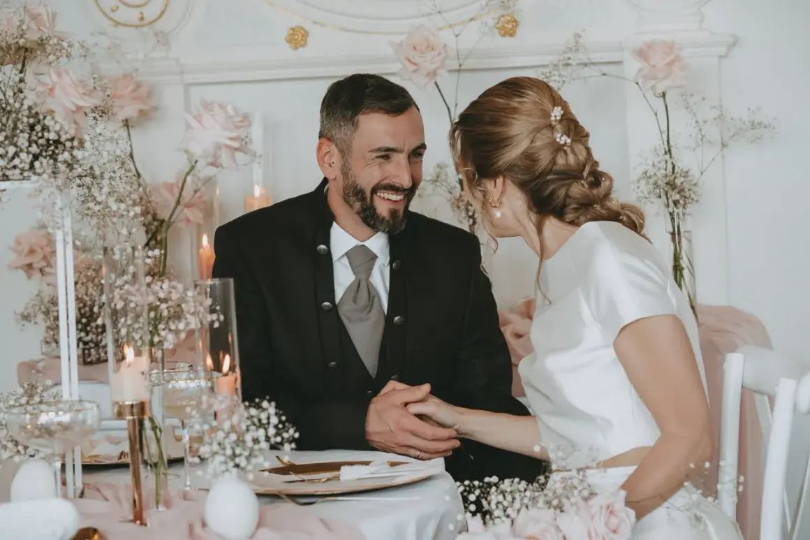 Ein glückliches Brautpaar bei ihrer Petersilienhochzeit, umgeben von zarten rosa Rosen und Schleierkraut. Sie halten Händchen und blicken sich liebevoll an.