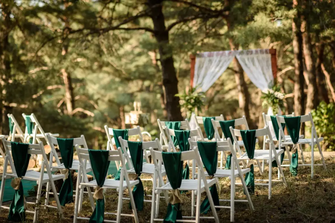 Reihen weißer Stühle mit grünen Schleifen schmücken den Weg zur Trauung. Eine romantische Hochzeit im Wald.