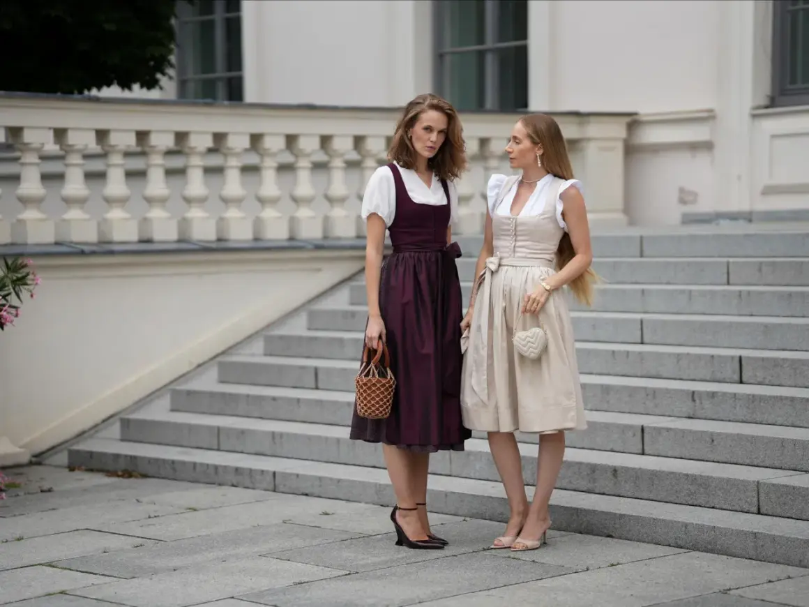 Zwei Frauen in eleganten Dirndl-Kleidern, ein moderner Dirndl-Trend, stehen auf einer Steintreppe vor einem Gebäude.