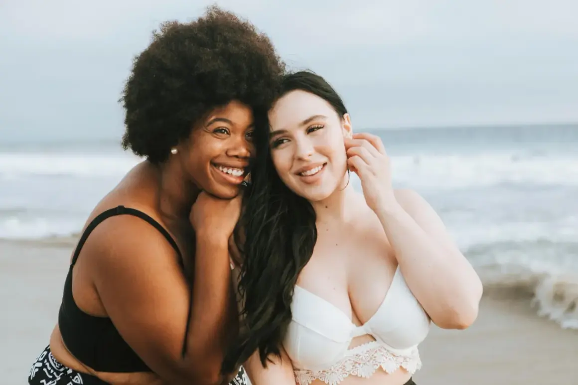 Zwei Frauen am Strand, eine mit Afro, die andere mit langen dunklen Haaren. Beide tragen Bikinis und lächeln. Ihre Brust und Bikinifigur sind gut sichtbar.
