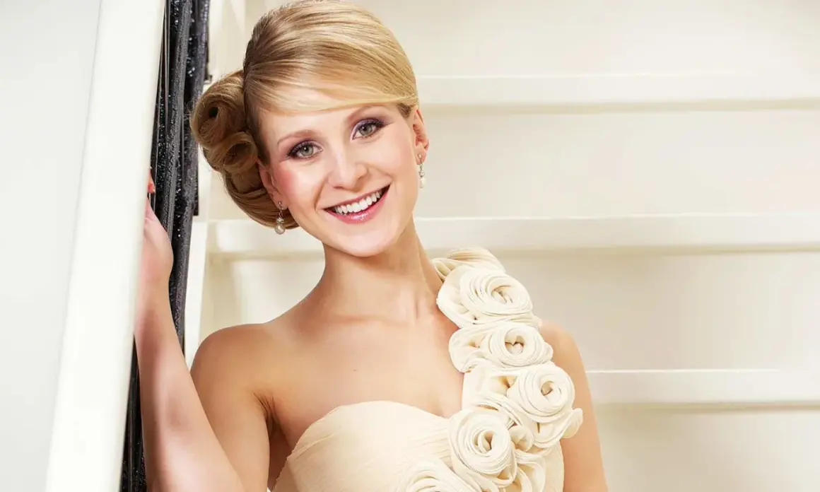 Lächelnde Frau in einem schulterfreien Kleid in Farbe ivory, verziert mit Stoffrosen. Sie lehnt an einem Geländer.