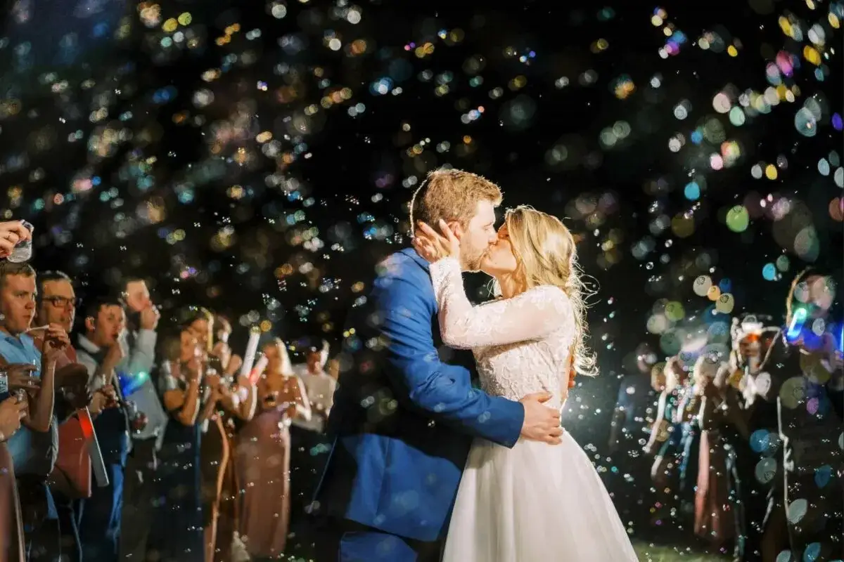 Brautpaar k&uuml;sst sich im Seifenblasenregen. G&auml;ste mit Lichtern im Hintergrund. Ein magischer Moment beim Auszug Hochzeit.