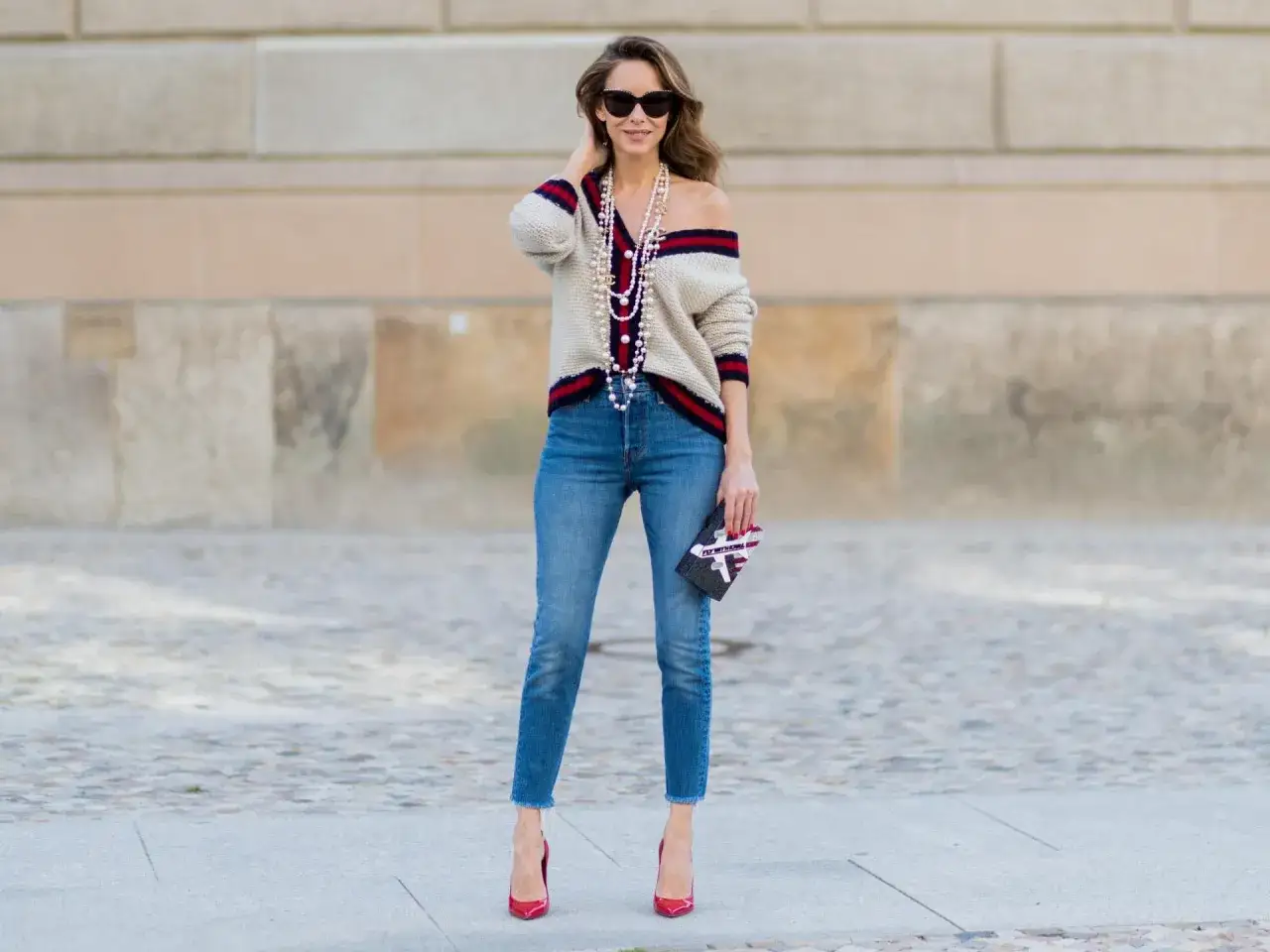 Moderne Damen tragen figurbetonte Jeans mit Fransen-Saum, dazu einen schulterfreien Cardigan und rote Pumps.